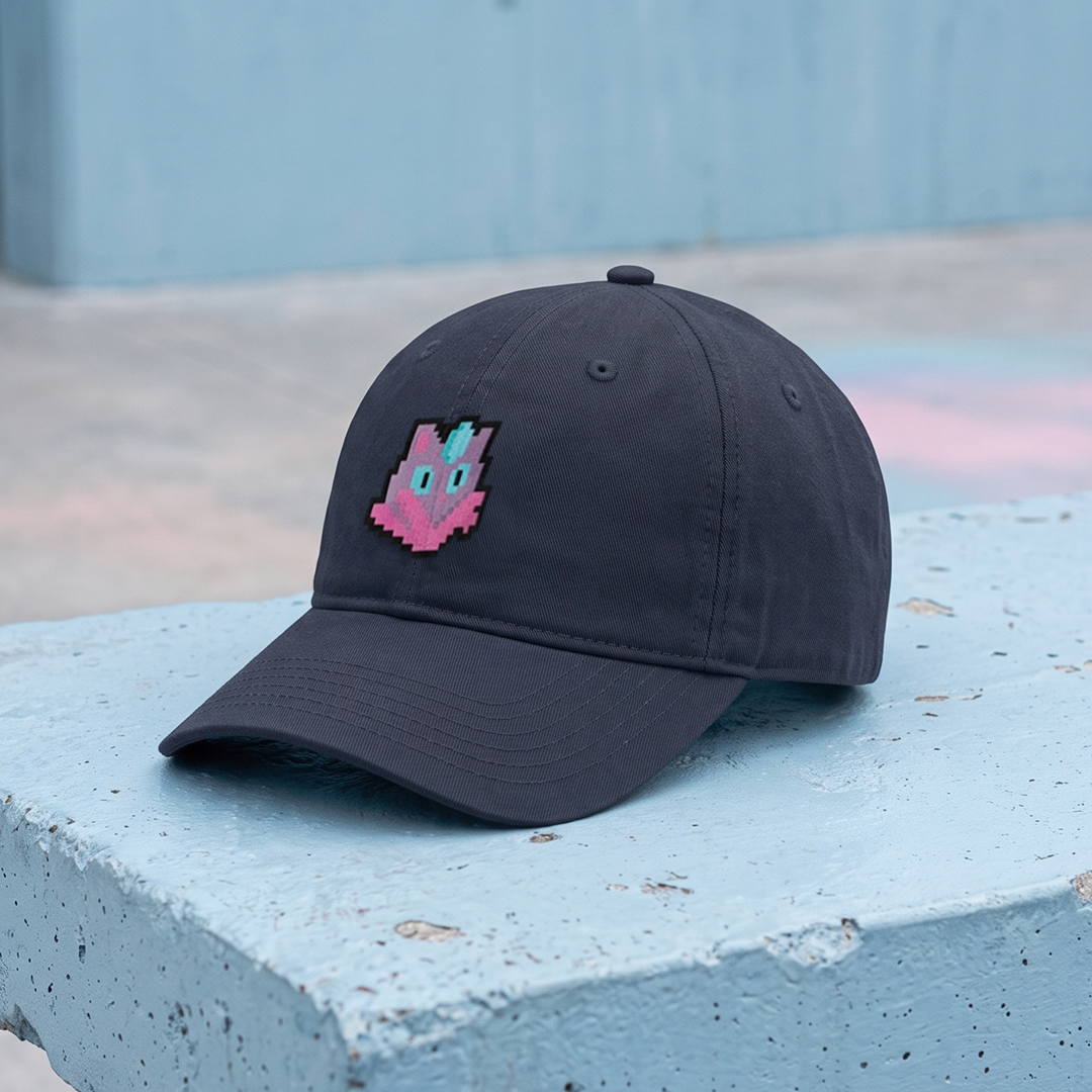 A dark charcoal gray baseball cap with a Amiko Legend character pixel art, resting on a light blue concrete ledge with a blurred blue-gray background.